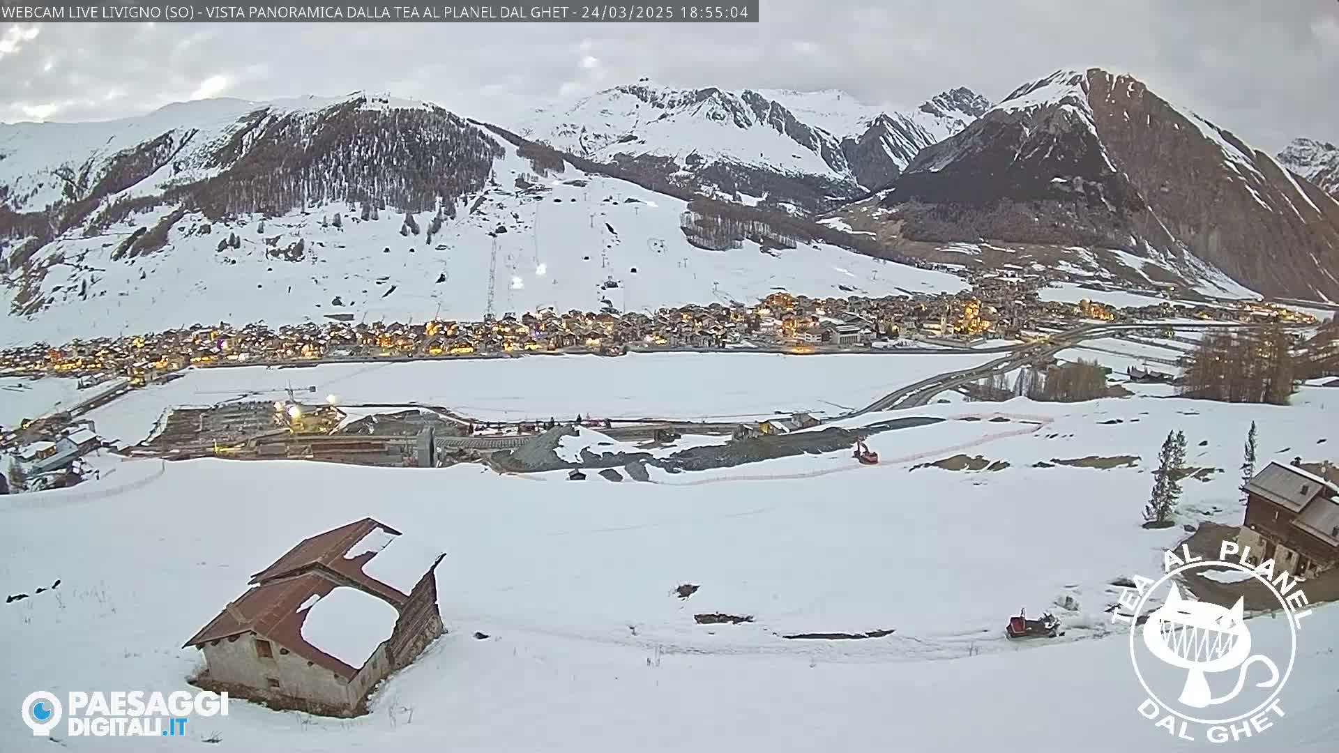 A snow-covered mountain village is nestled in a valley beneath snow-covered mountains, with a partly cloudy sky.