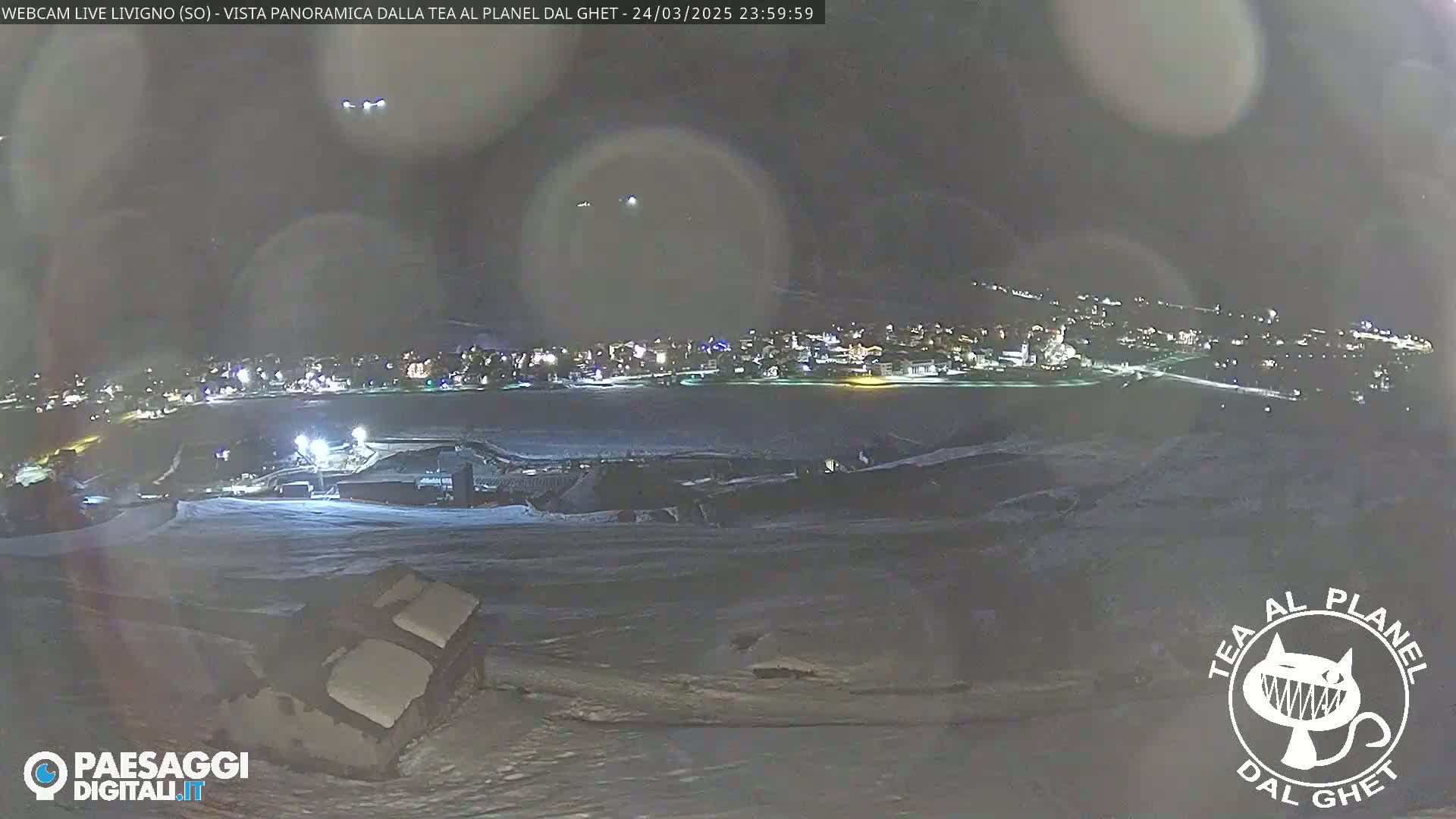 A nighttime, panoramic view of a snow-covered landscape shows a town in the distance with its lights on, under a dark, somewhat hazy sky.