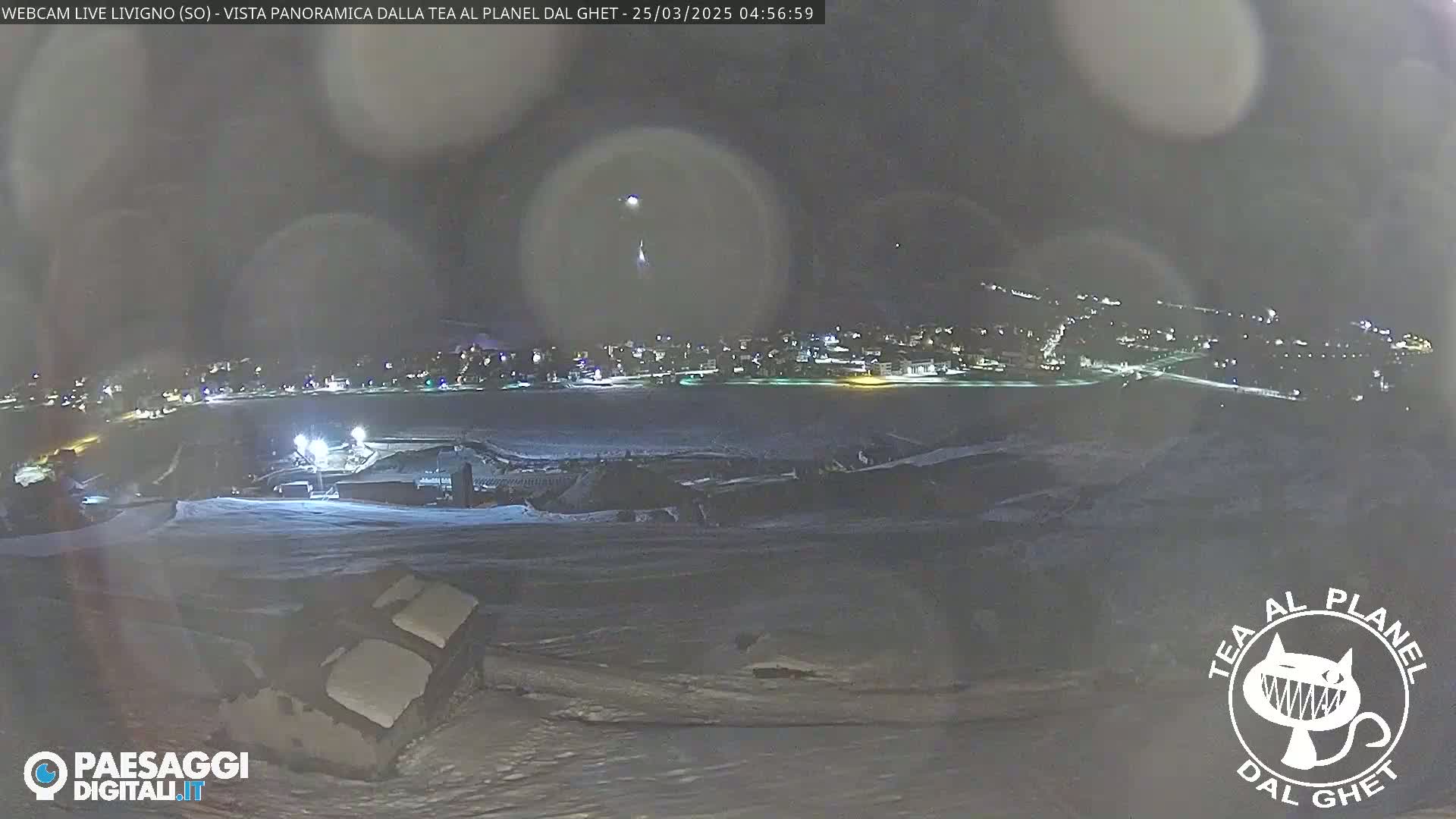 A nighttime, snowy landscape shows a town in the distance illuminated by lights, with a partially visible structure in the foreground under a dark, possibly cloudy sky.