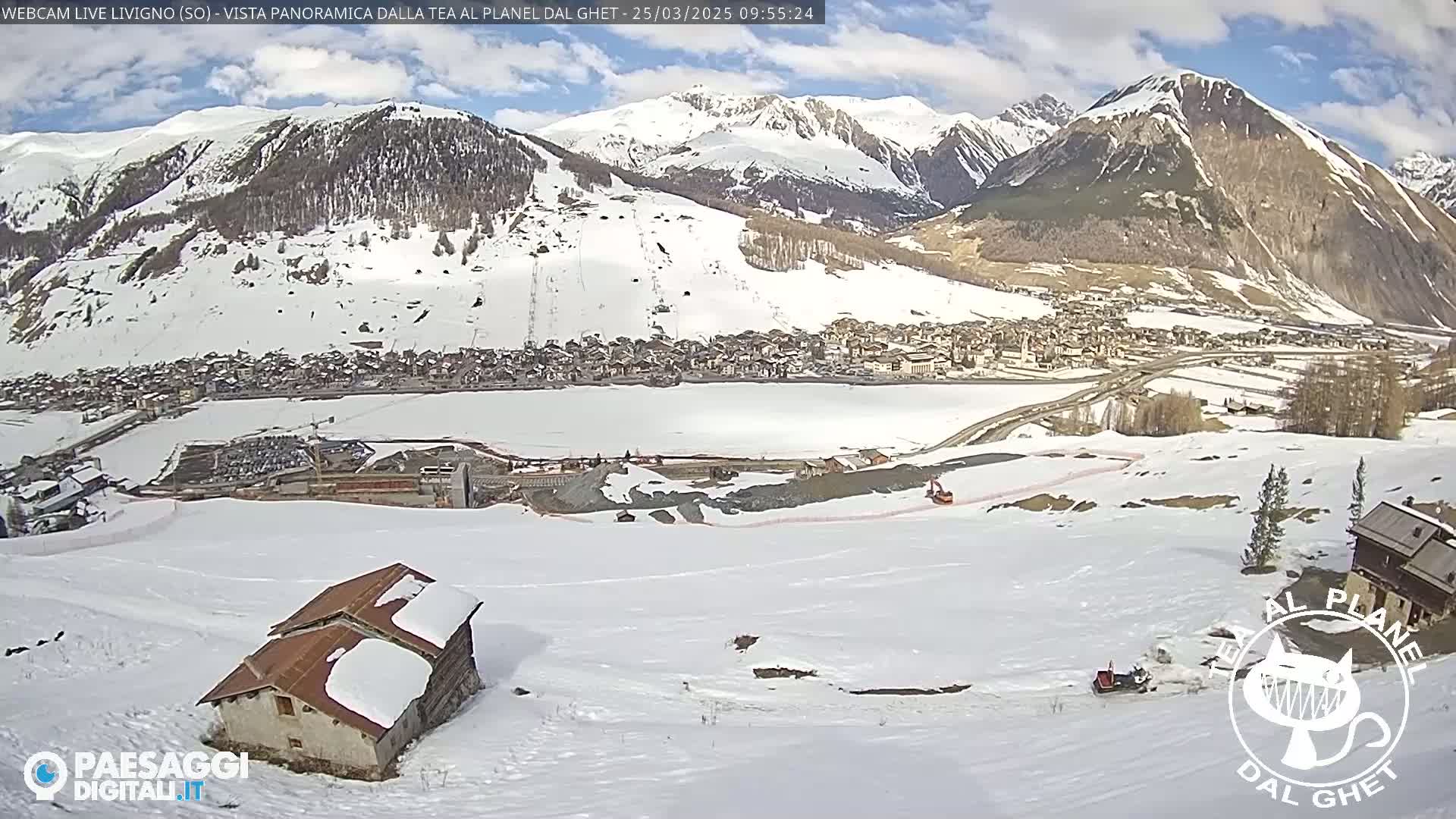 A snow-covered mountain village is nestled below a range of snow-capped mountains under a partly sunny sky.