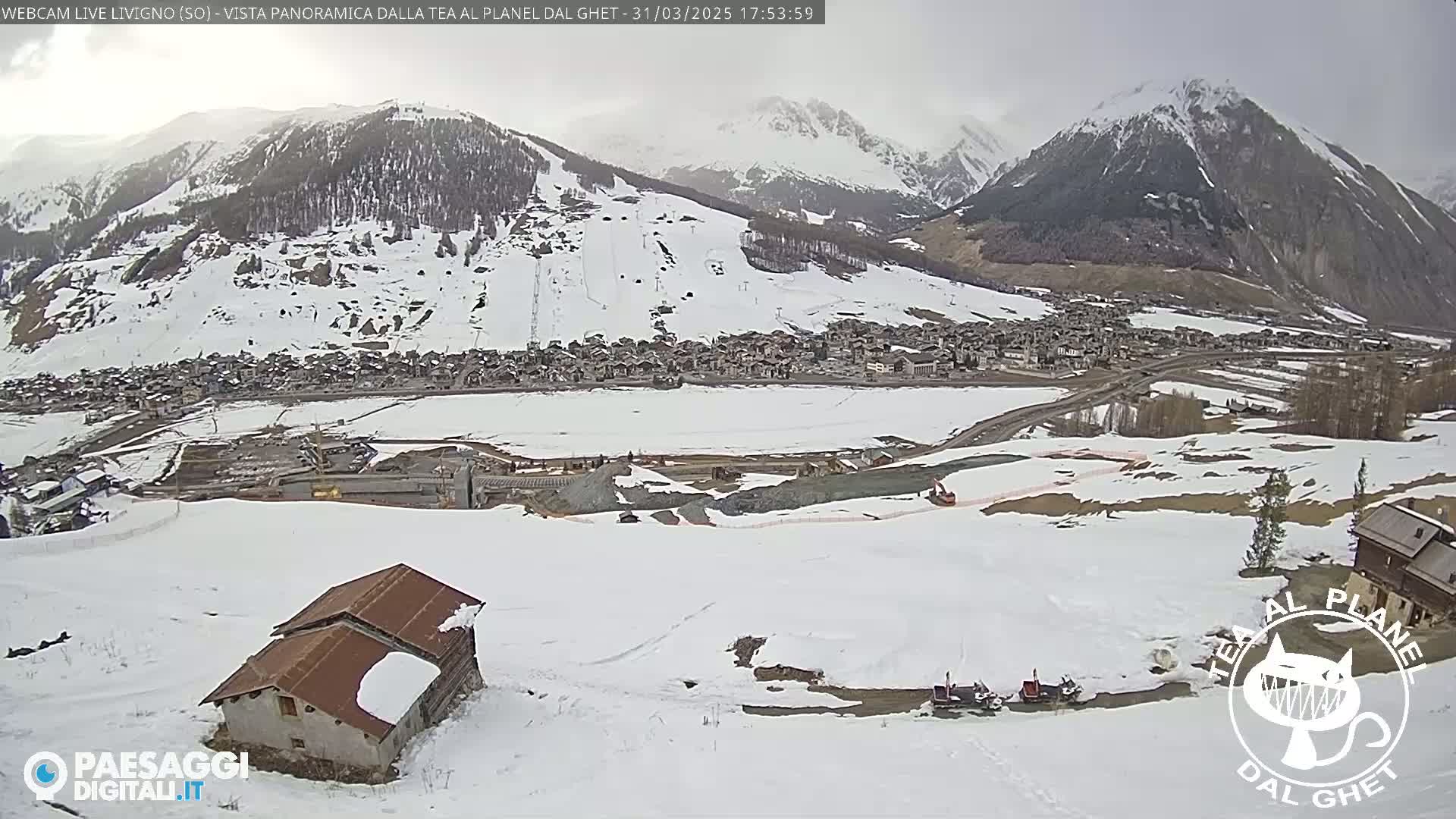 A snow-covered mountain village is viewed from a high vantage point on a cloudy day, with a small building and some snowmobiles visible in the foreground.