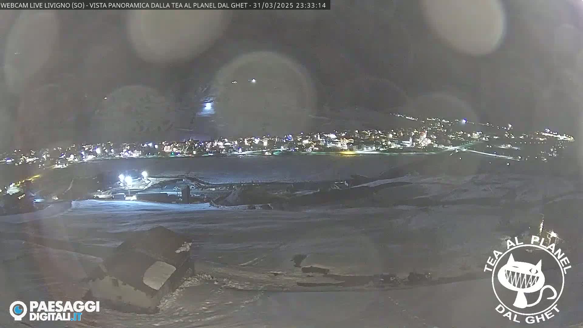A nighttime, panoramic view of a snow-covered town illuminated by lights, seen from a higher vantage point under a dark, hazy sky.