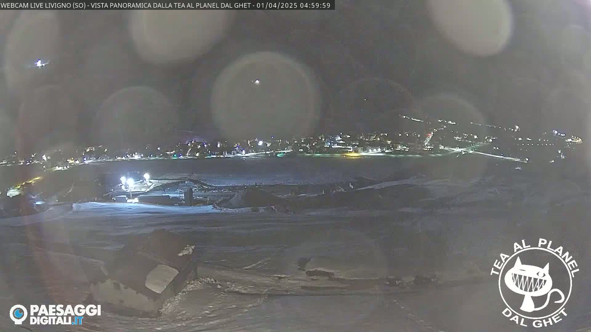A nighttime, snowy landscape shows a distant, illuminated town under a dark sky with some lens flares.