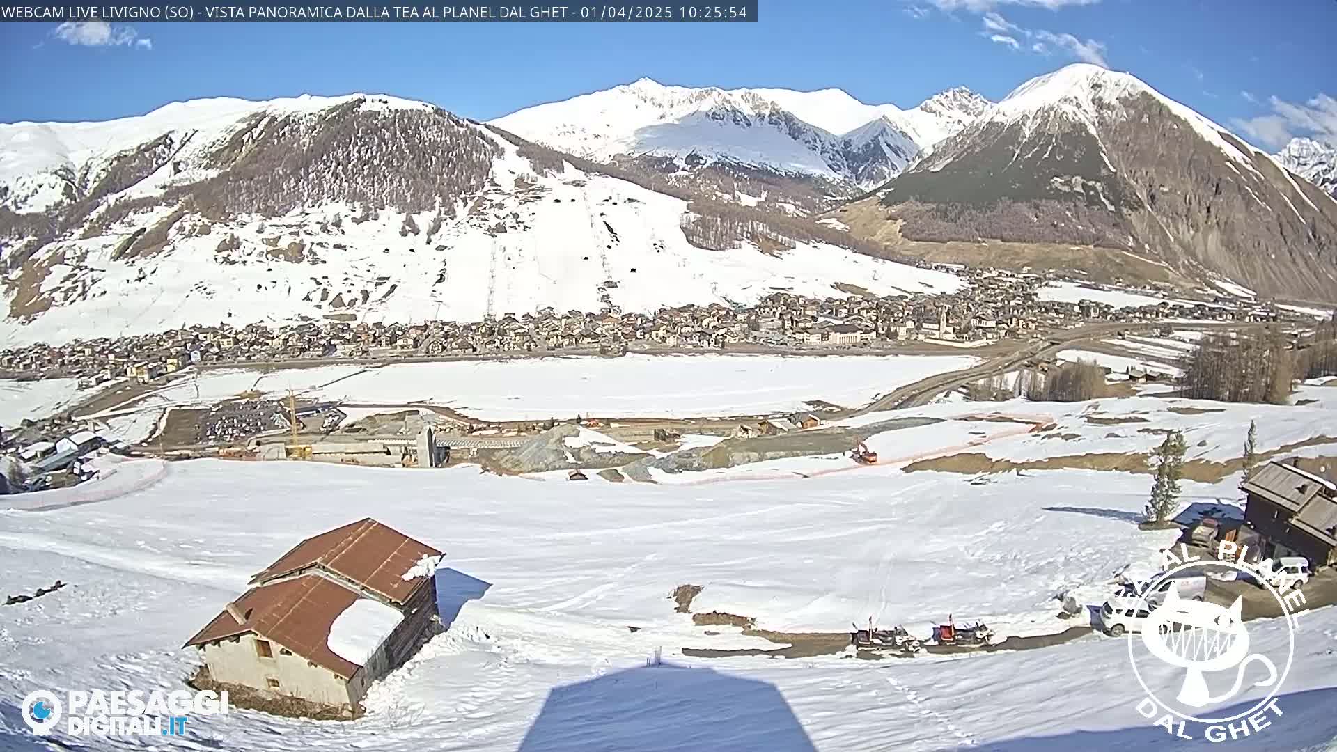 A snow-covered mountain village is nestled in a valley beneath snow-capped mountains under a mostly sunny sky.