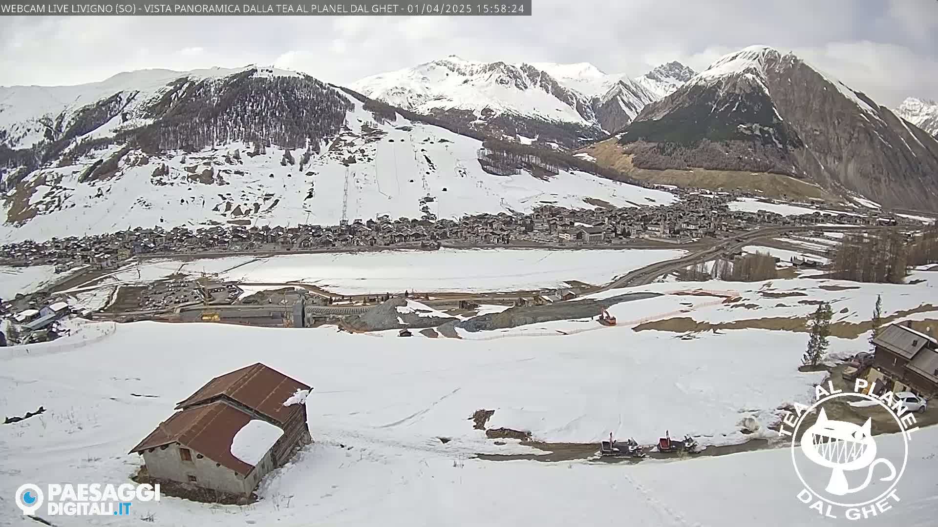 A snow-covered mountain valley village is viewed from a high vantage point on a partly cloudy day, with a small building in the foreground.
