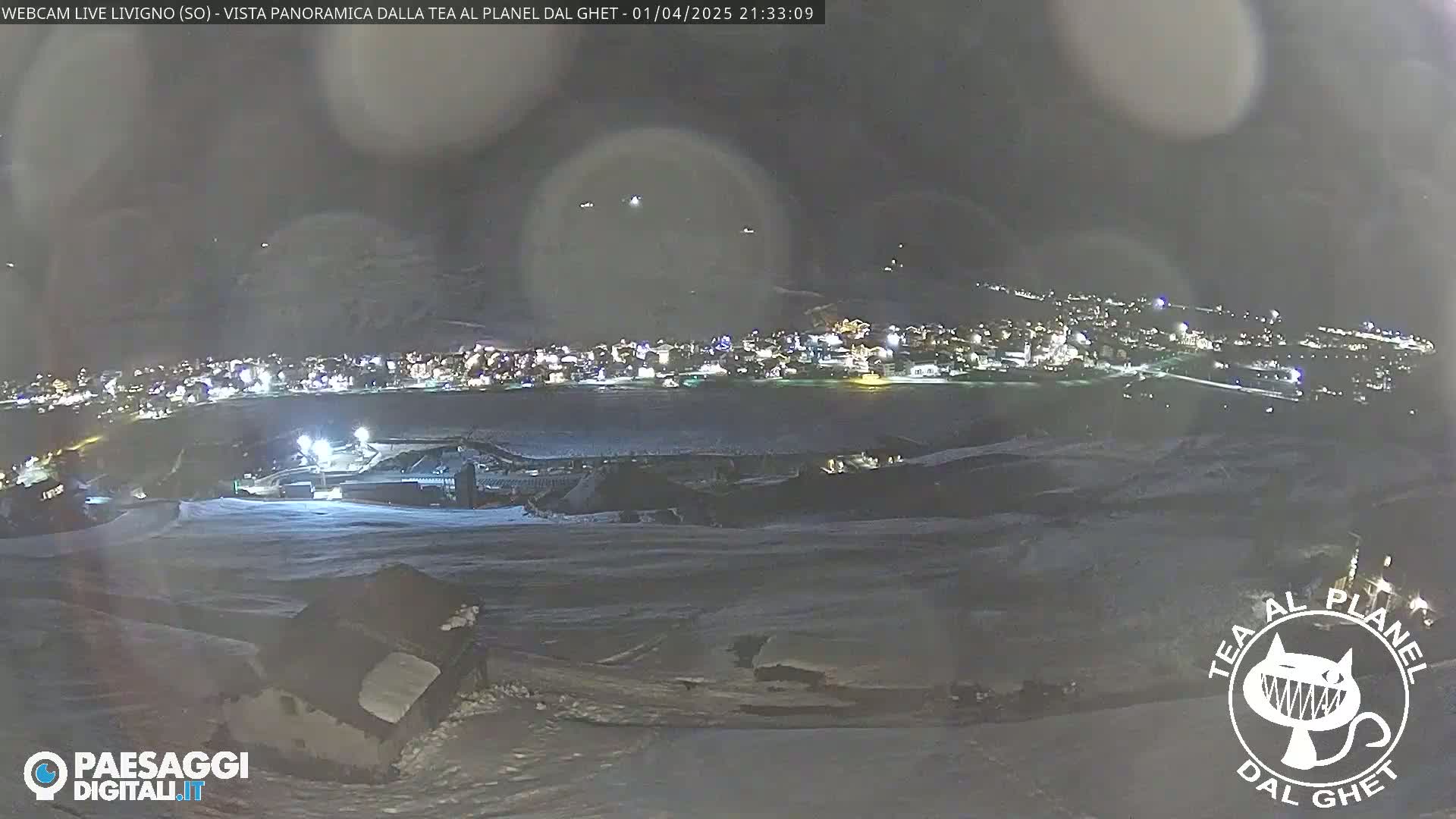 A nighttime, snow-covered landscape shows a distant town illuminated with lights, while the foreground reveals a sparsely populated snowy area.