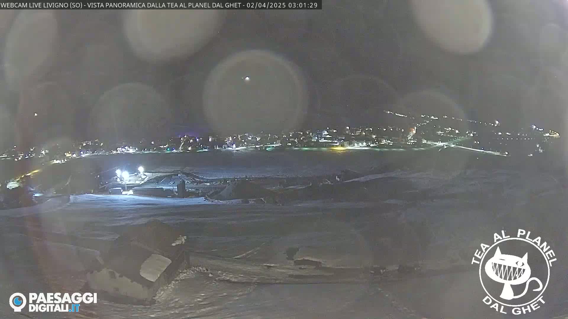 A nighttime panoramic view of a snow-covered landscape shows a town with illuminated buildings in the distance under a dark sky with faint lights.