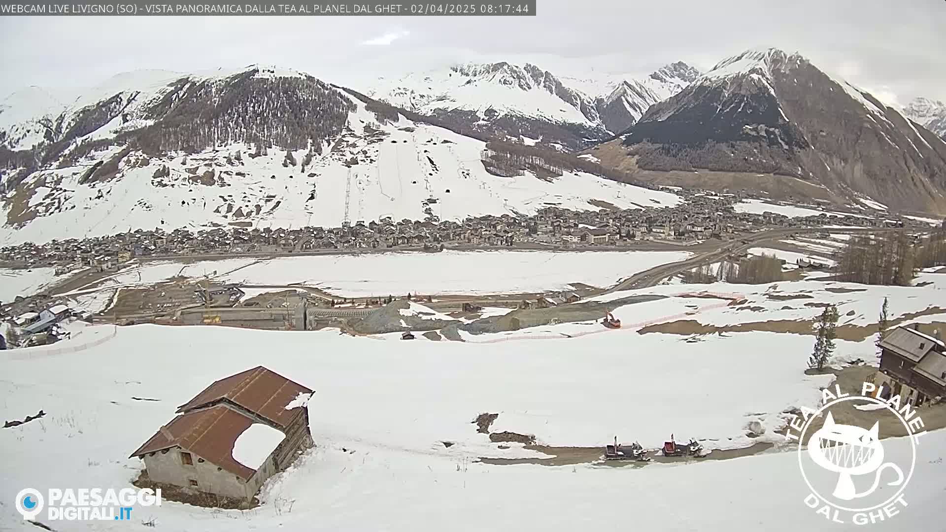A snow-covered mountain village is seen from a high vantage point on a cloudy day, with snow-covered slopes and a small building in the foreground.