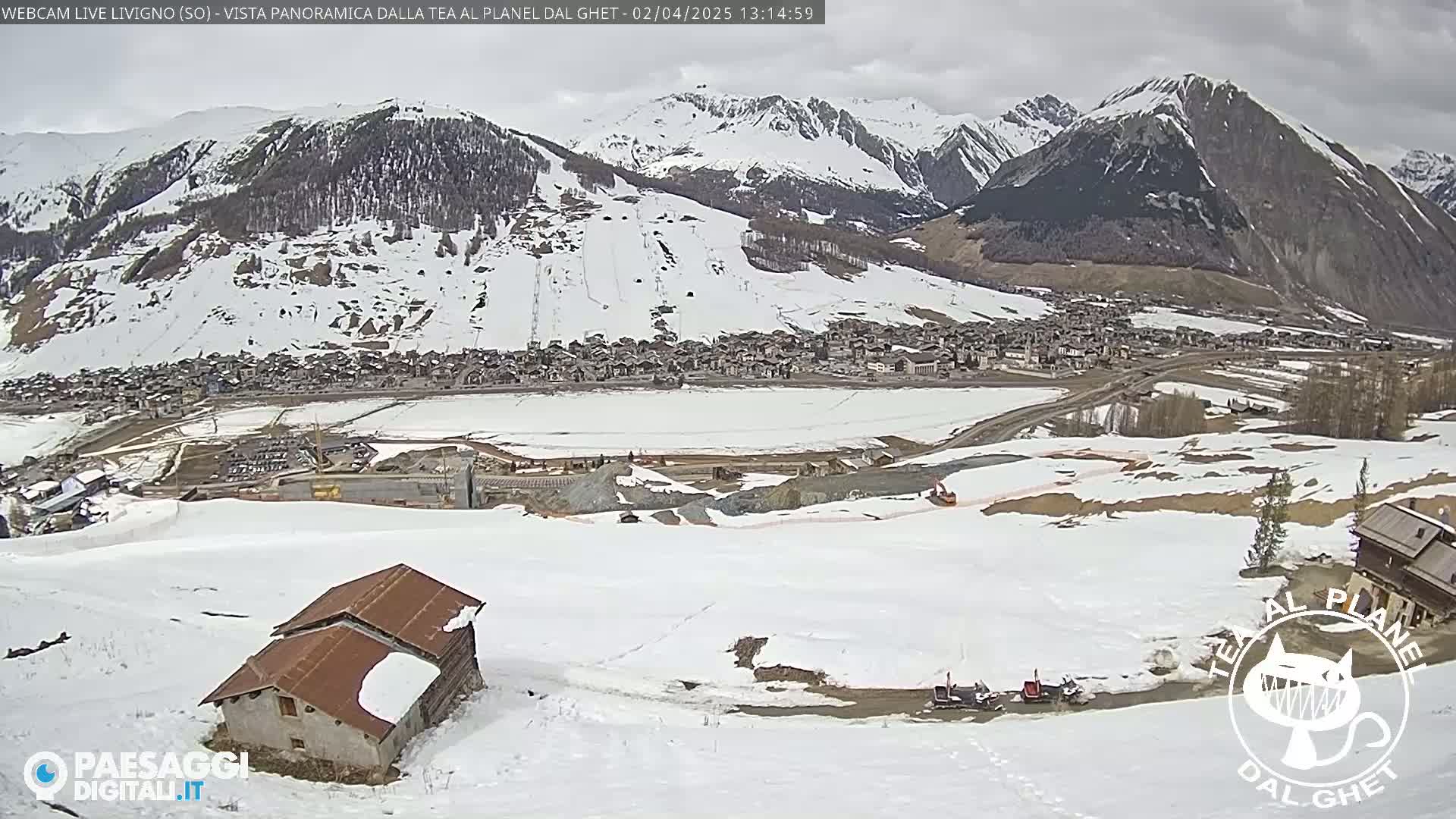 A snow-covered mountain village is visible beneath a cloudy sky, with a small building and some snowmobiles in the foreground.