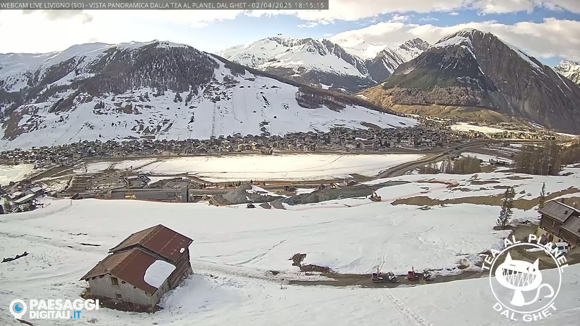 A snow-covered mountain village is nestled at the base of snow-capped mountains under a mostly sunny sky.