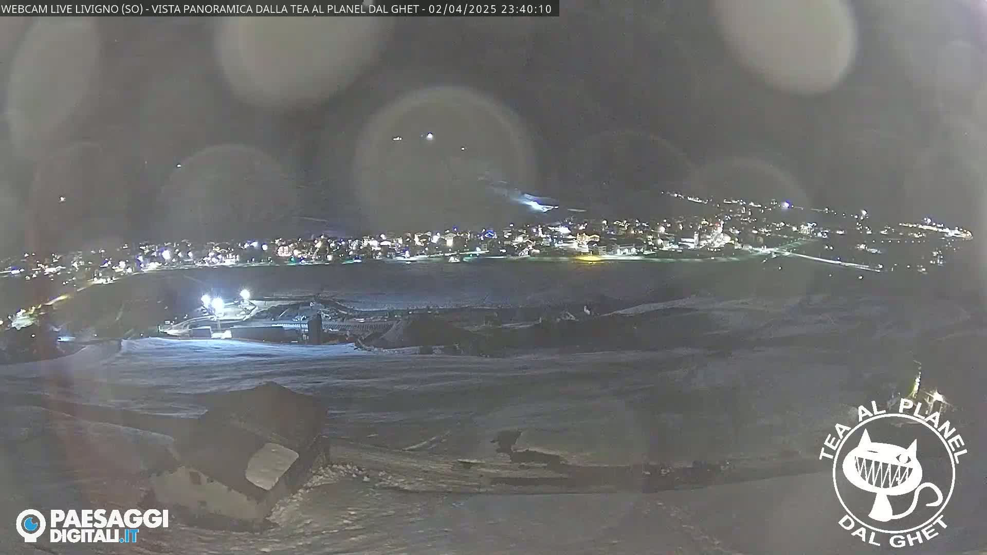 A nighttime panoramic view of a snow-covered landscape showing a town's lights in the distance under a dark, slightly hazy sky.