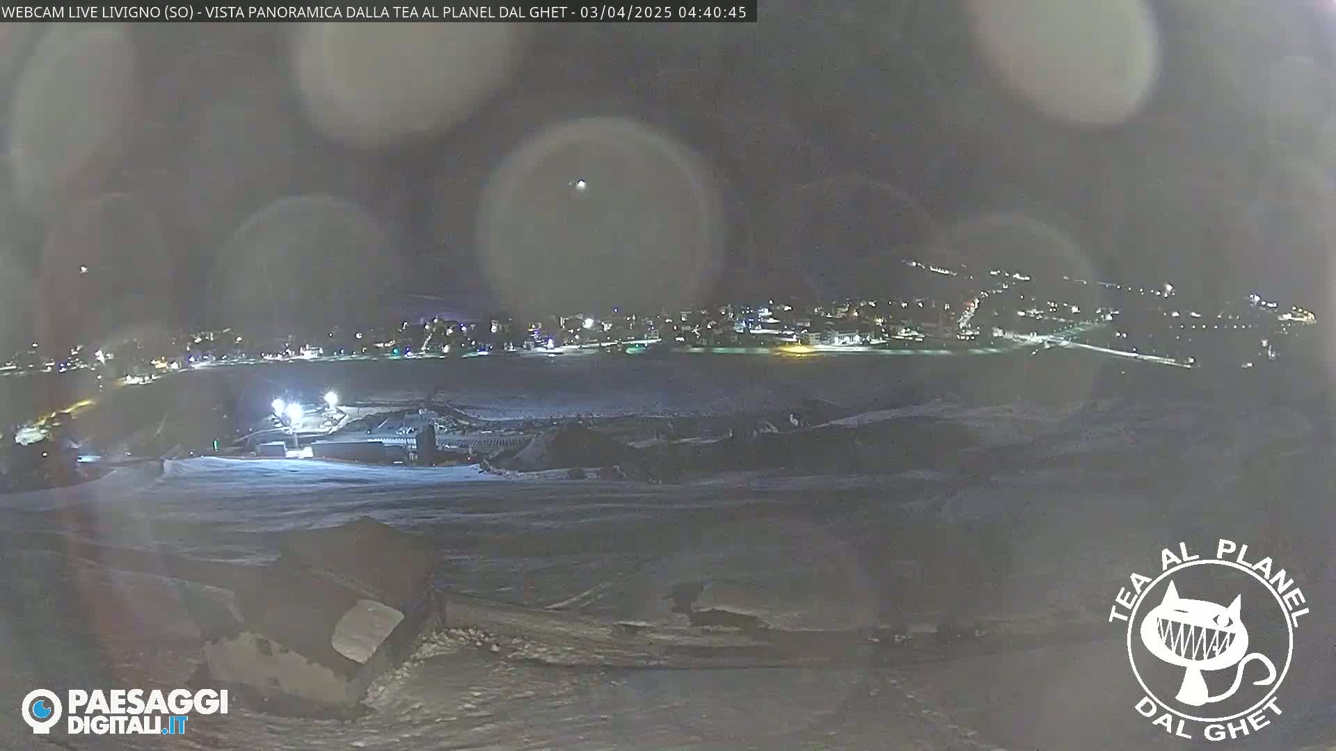 A nighttime, snowy panoramic view of a town illuminated by lights, with hazy atmospheric conditions.