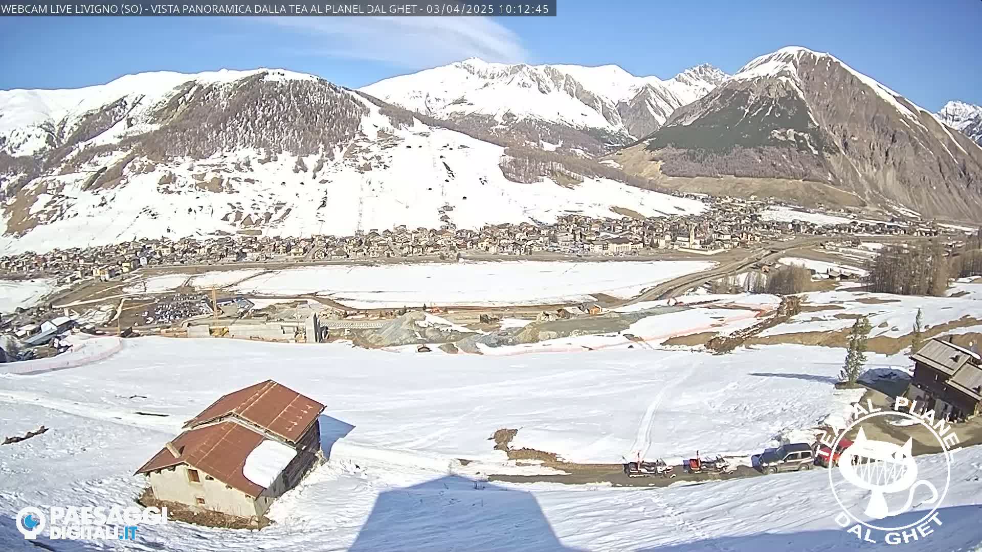 A snow-covered mountain valley town is seen from a high vantage point on a sunny day, with a small building and some vehicles visible in the foreground.