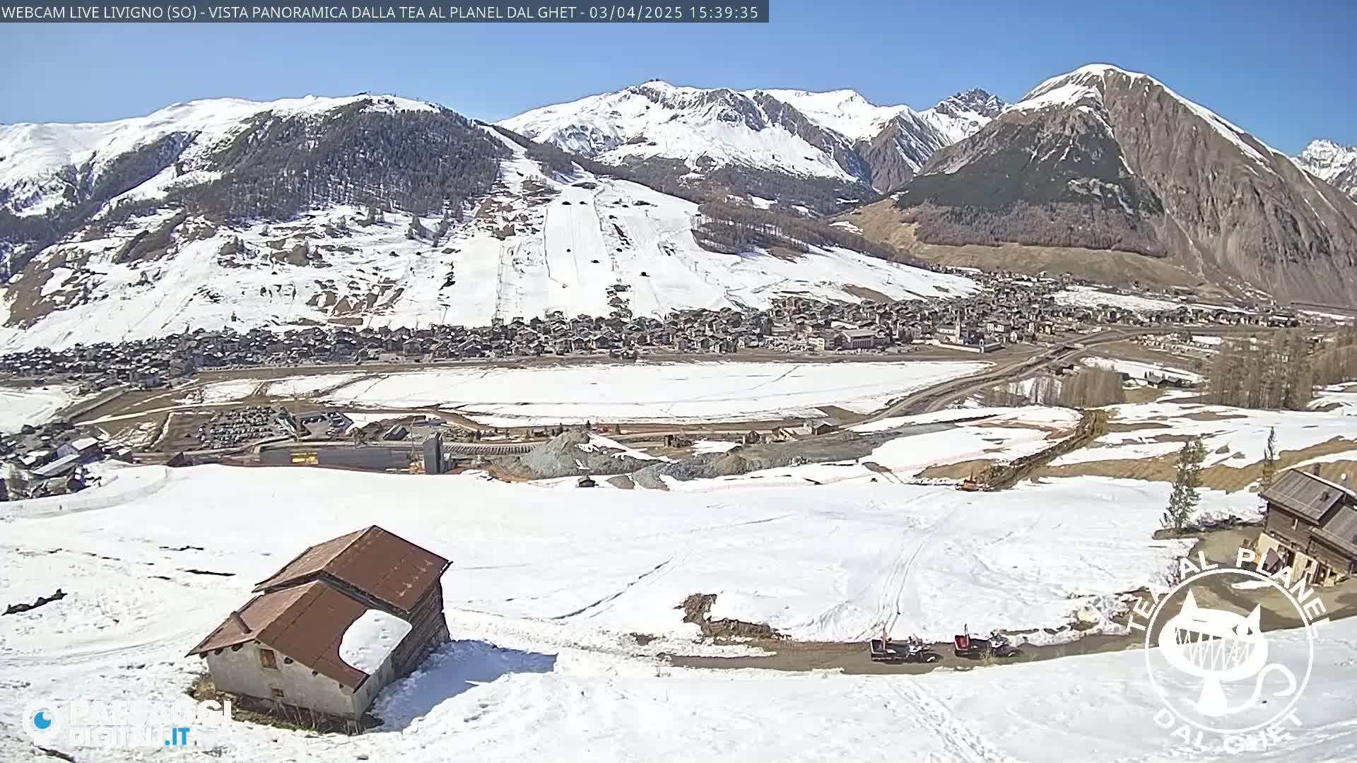 A snow-covered mountain valley village is seen under a sunny sky, with a dilapidated building and some vehicles visible in the foreground.