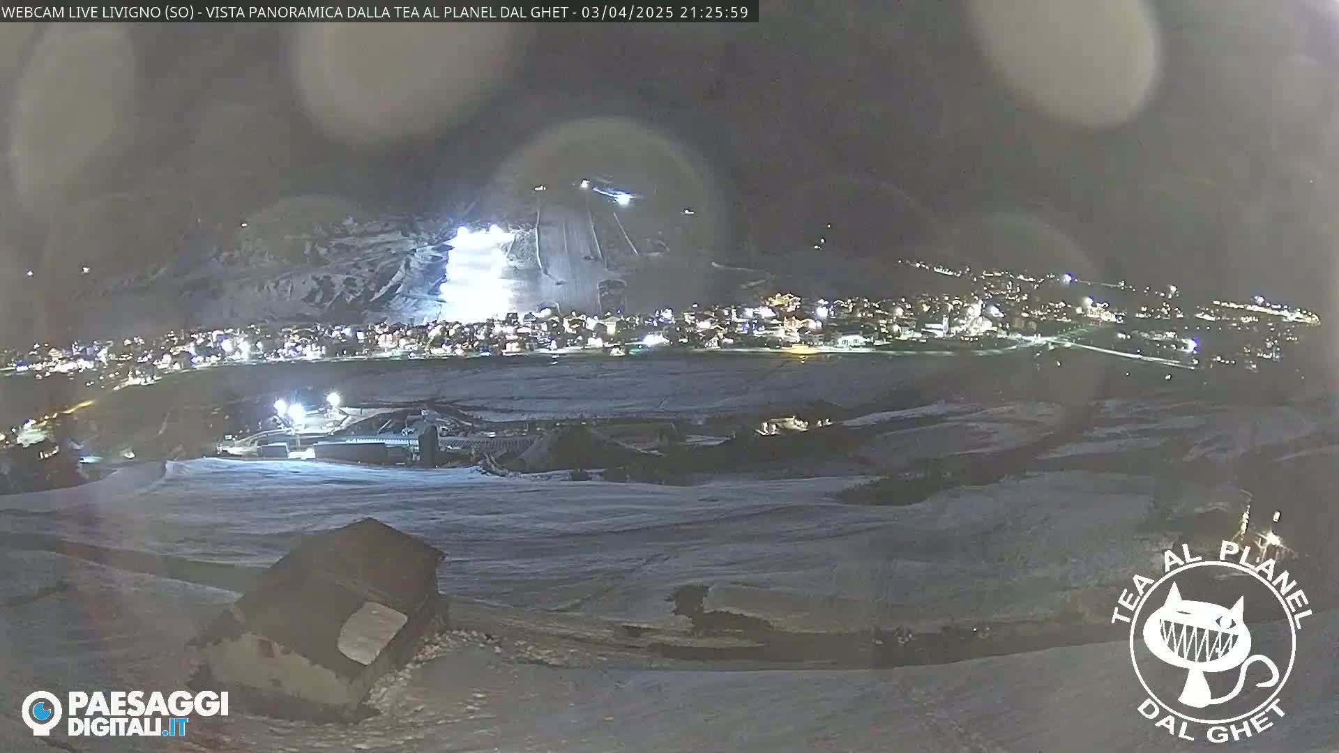 A nighttime, snow-covered landscape shows a town brightly lit below, with illuminated ski slopes above, under a dark, cloudy sky.
