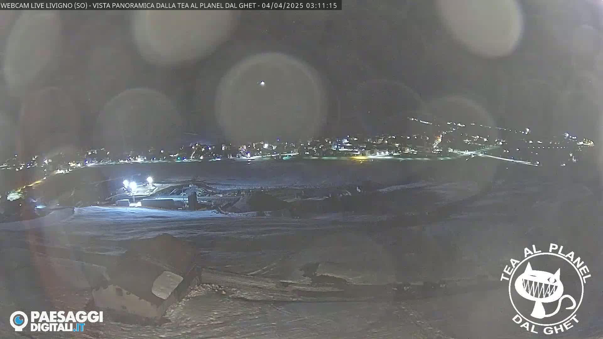 A nighttime, snowy scene shows a town illuminated in the distance, viewed from a snow-covered vantage point with hazy atmospheric conditions.