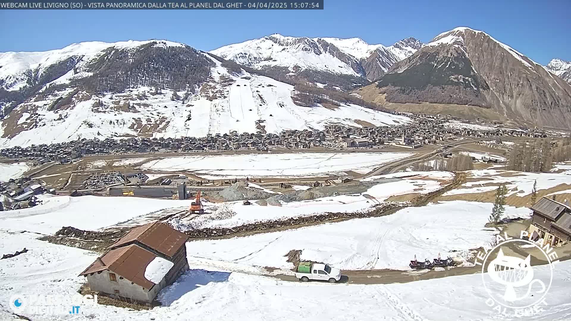 A sunny, snow-covered mountain landscape reveals a village nestled in a valley, with construction activity and vehicles visible on a partially snow-covered road below.