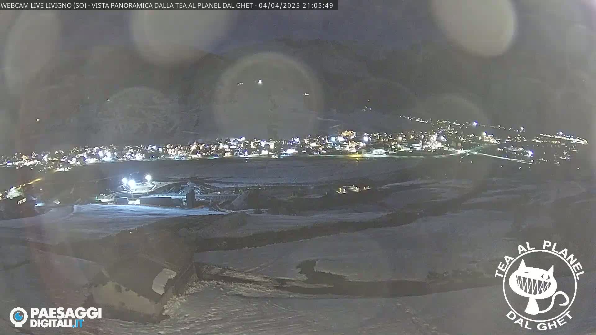 A nighttime, snowy view of a village nestled in a valley, illuminated by lights, under a hazy sky.
