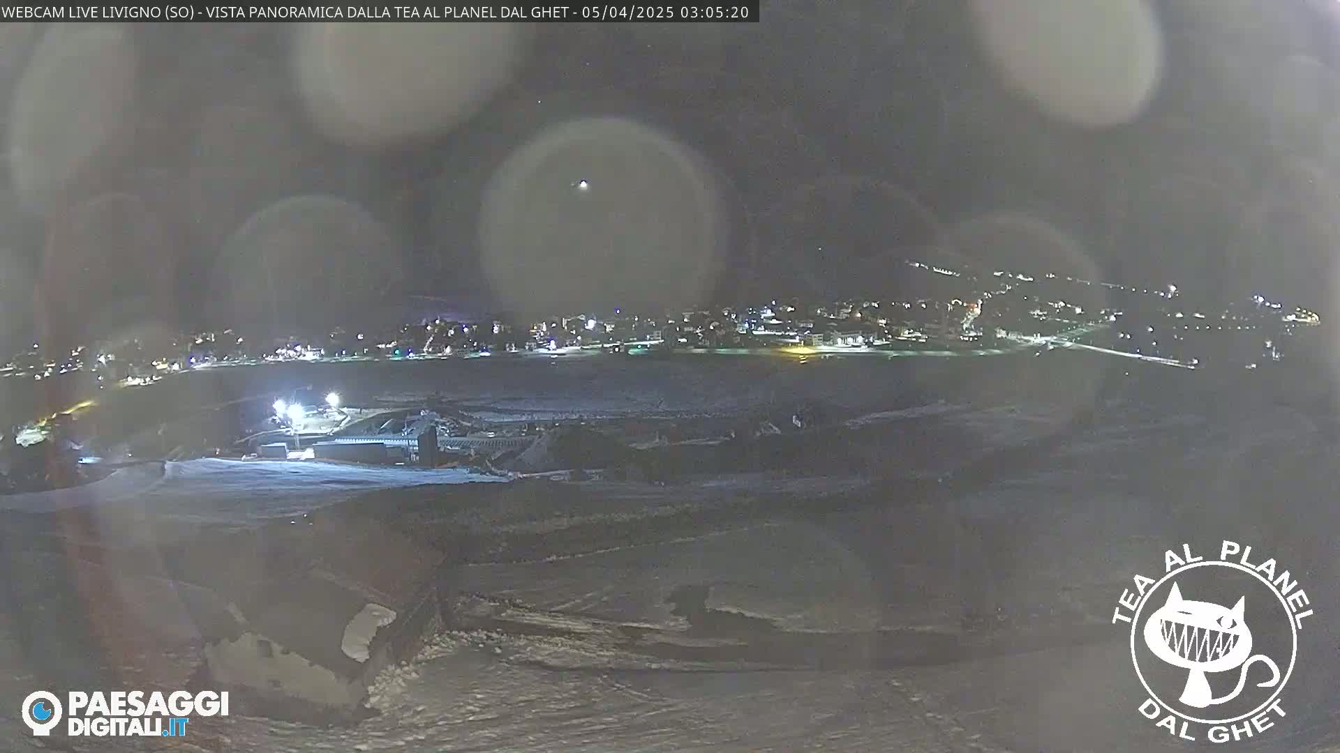 A nighttime, snow-covered landscape shows a town with illuminated buildings in the distance, under a dark, slightly hazy sky.