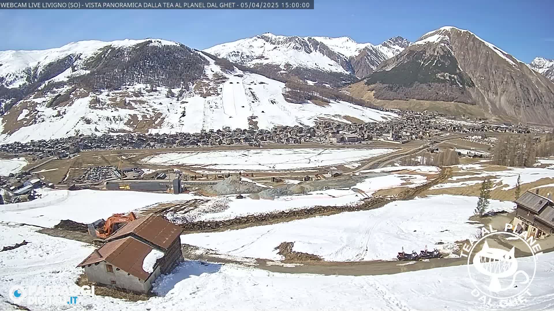 A snow-covered mountain valley town is viewed from a high vantage point on a sunny day, with construction visible in the foreground and snow-covered ski slopes in the background.