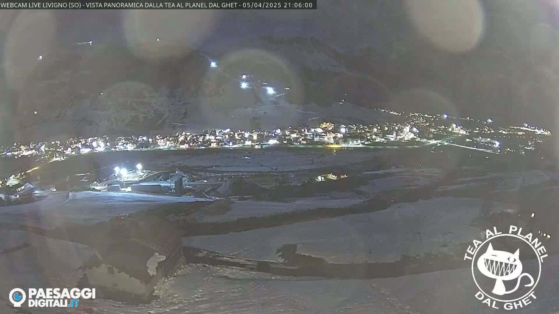 A nighttime, snow-covered landscape shows a town illuminated below a dark mountain, with faint lights indicating activity on a ski slope.