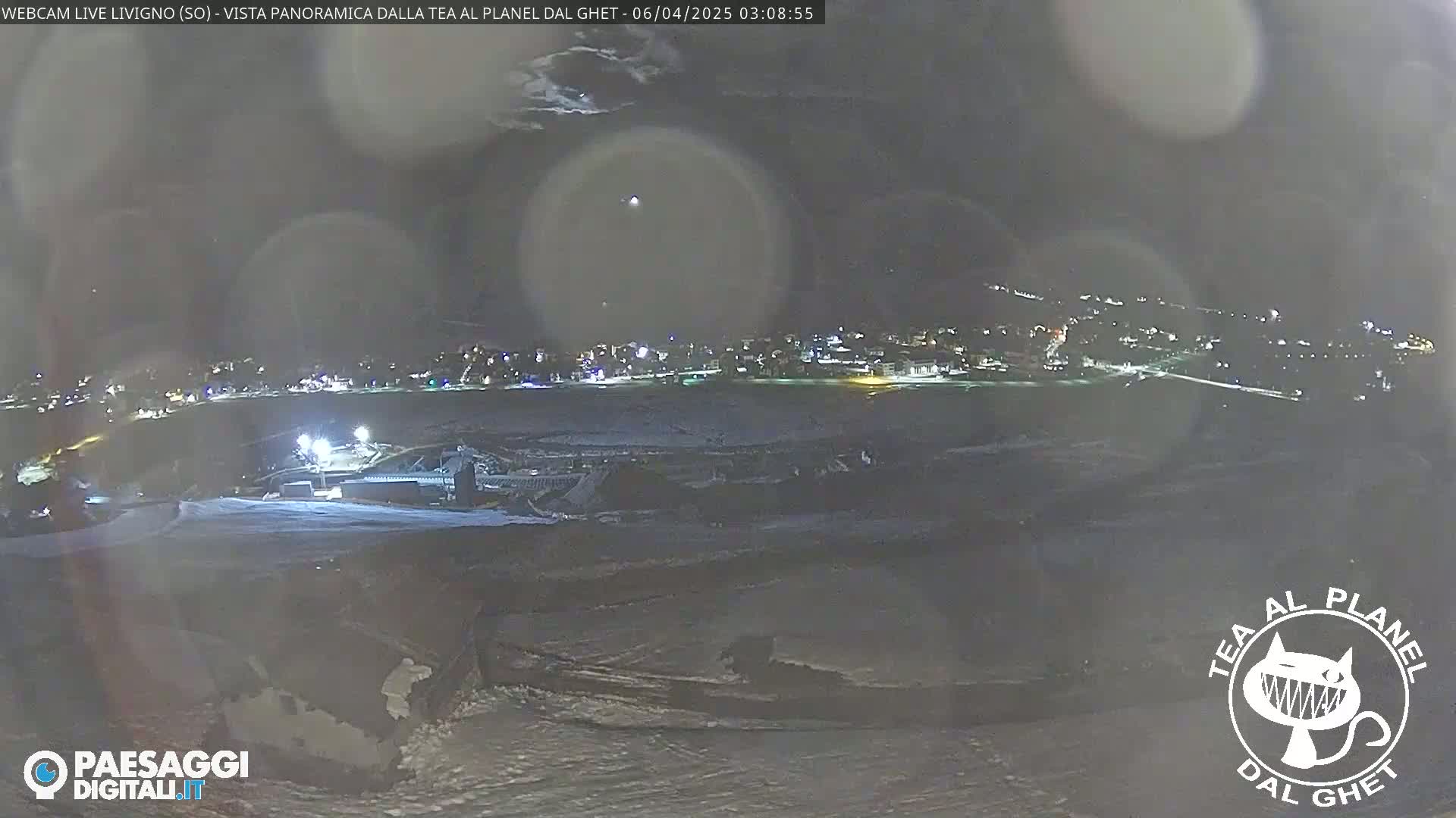A nighttime, panoramic view of a snow-covered landscape with a town illuminated in the distance under a dark, possibly cloudy sky.
