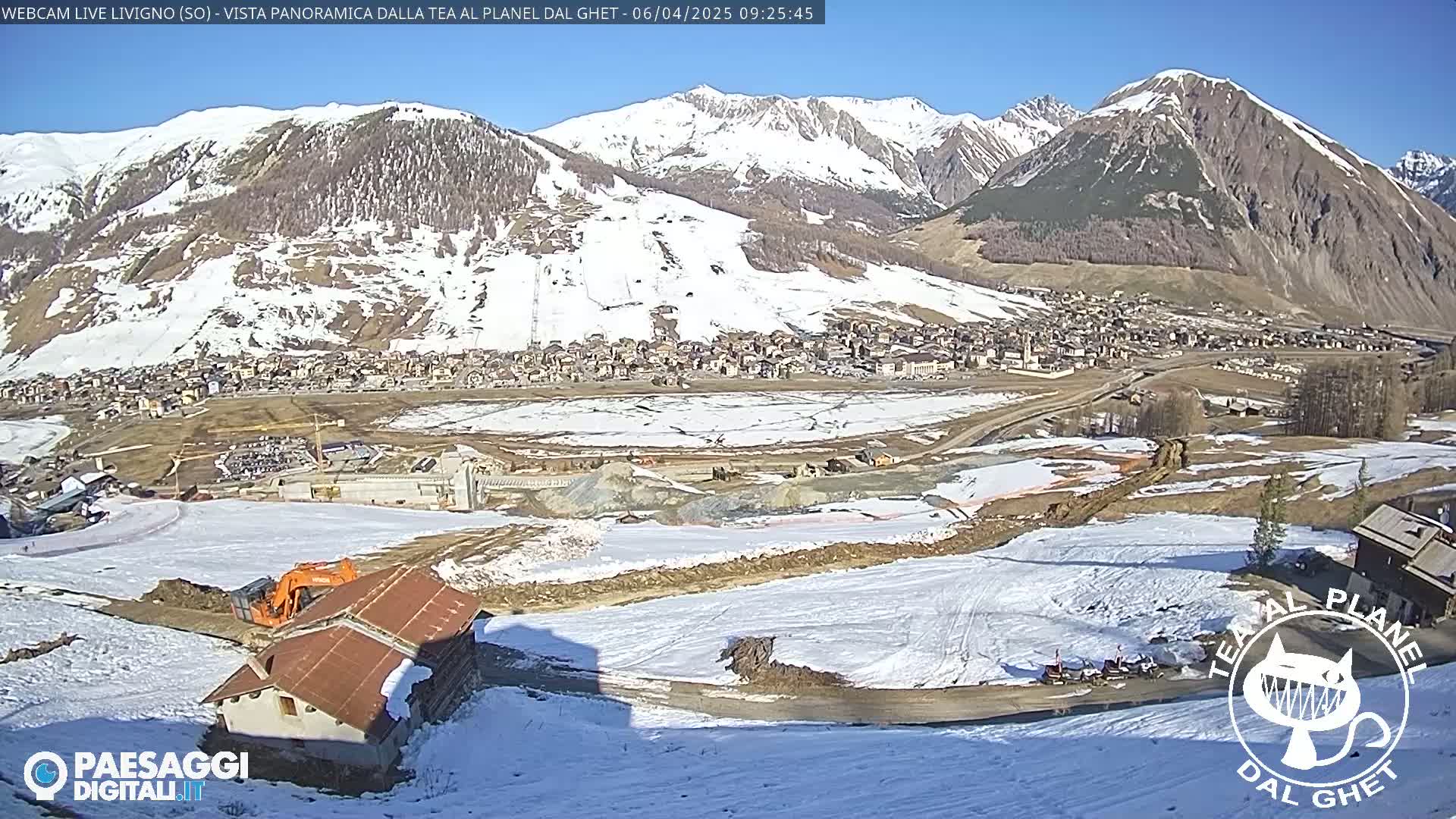 A sunny mountain village scene with snow-covered hills and construction activity in the foreground.