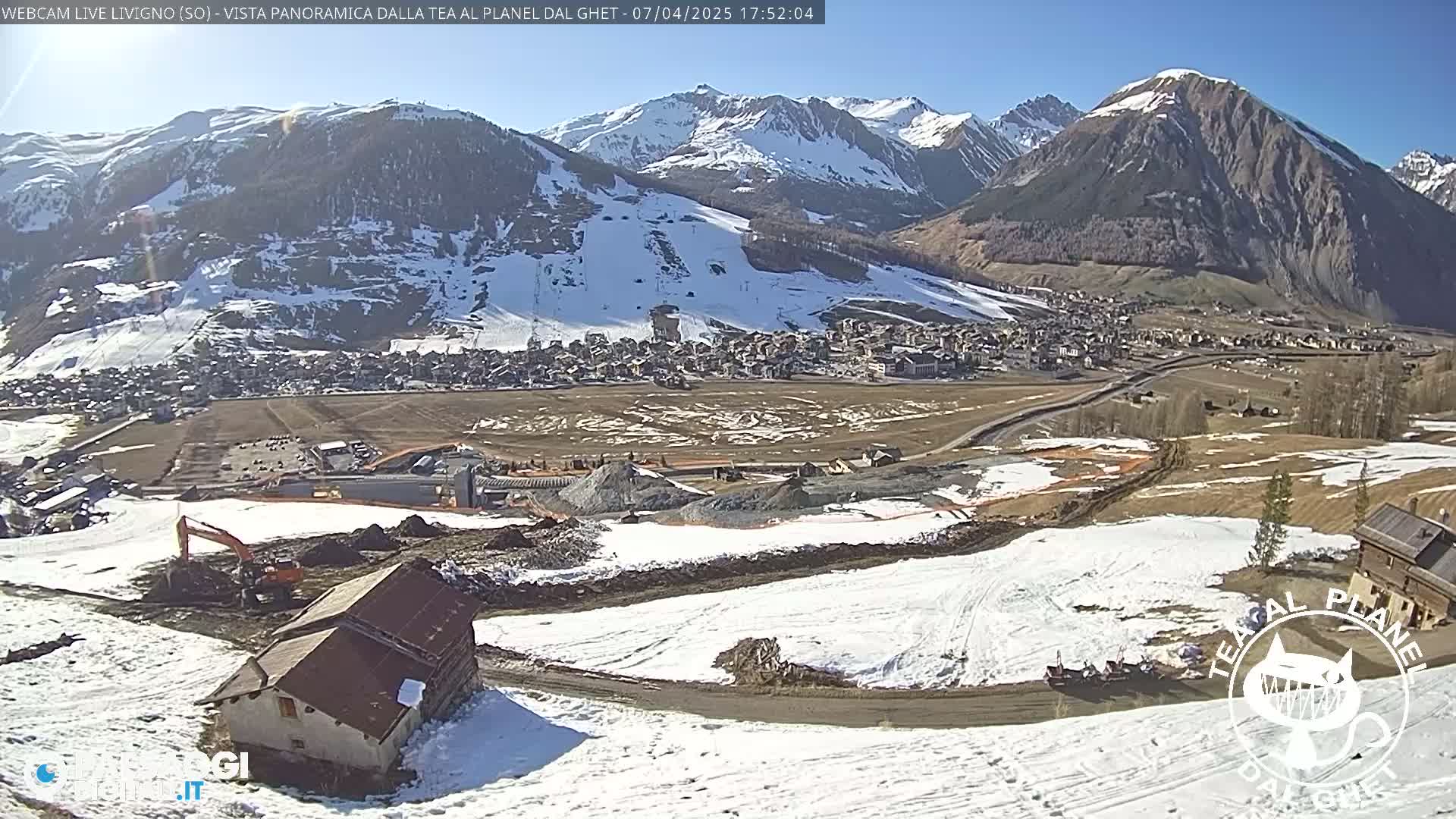 A sunny mountain village sits below snow-covered slopes and mountains, with construction activity visible in the foreground.