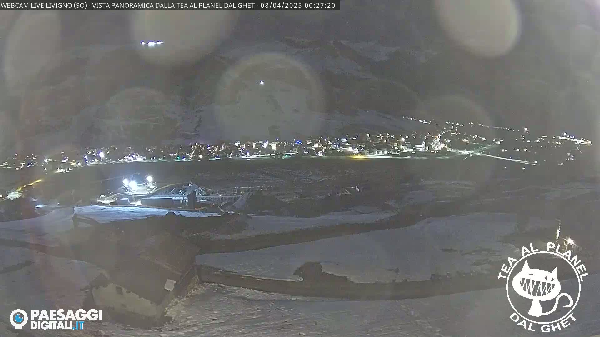 A nighttime, snow-covered landscape shows a town illuminated below, with a hazy sky above.