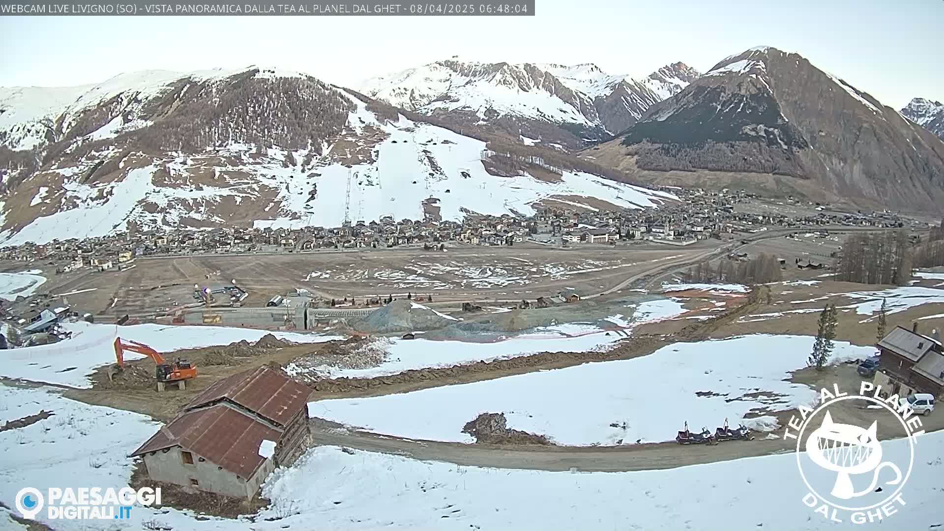 A partially snow-covered mountain village is viewed from a hilltop, with construction underway in the foreground and an orange excavator visible; some snow is present on the ground.