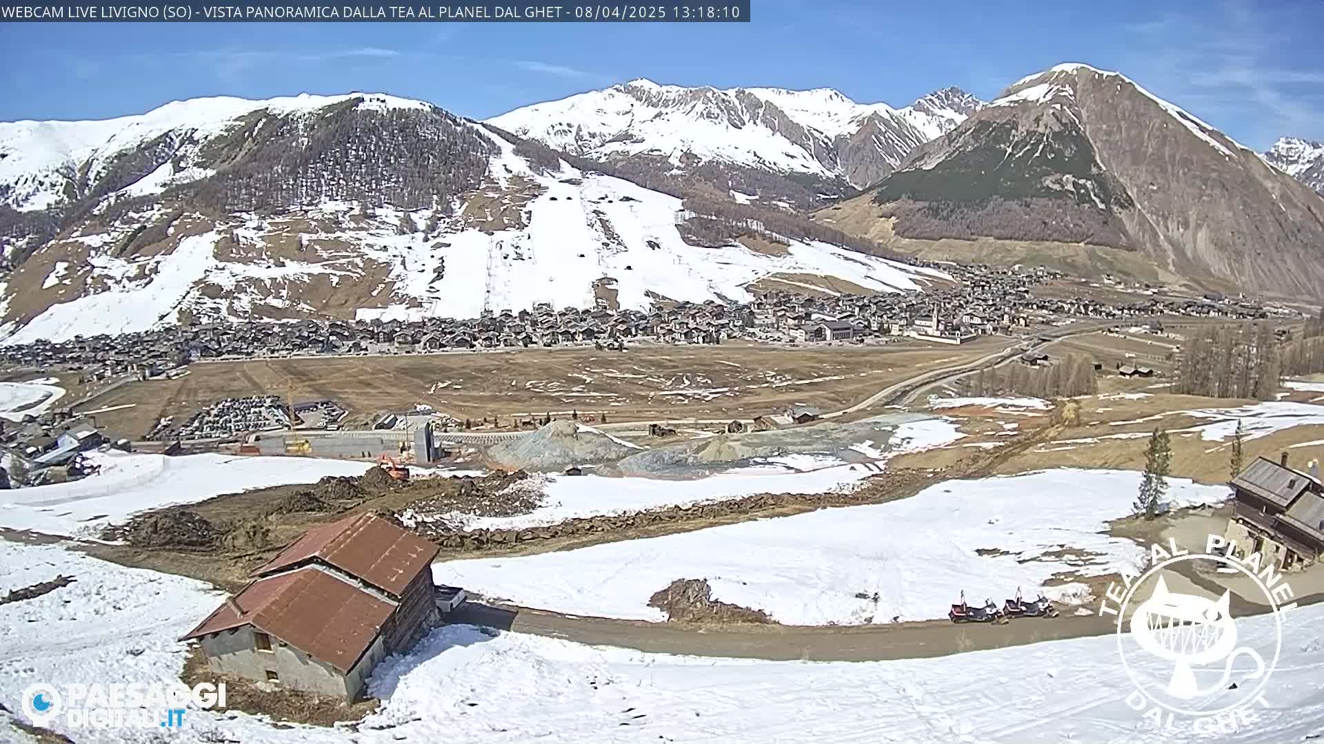 A mountain village nestled in a valley with snow-covered mountains in the background on a sunny day.