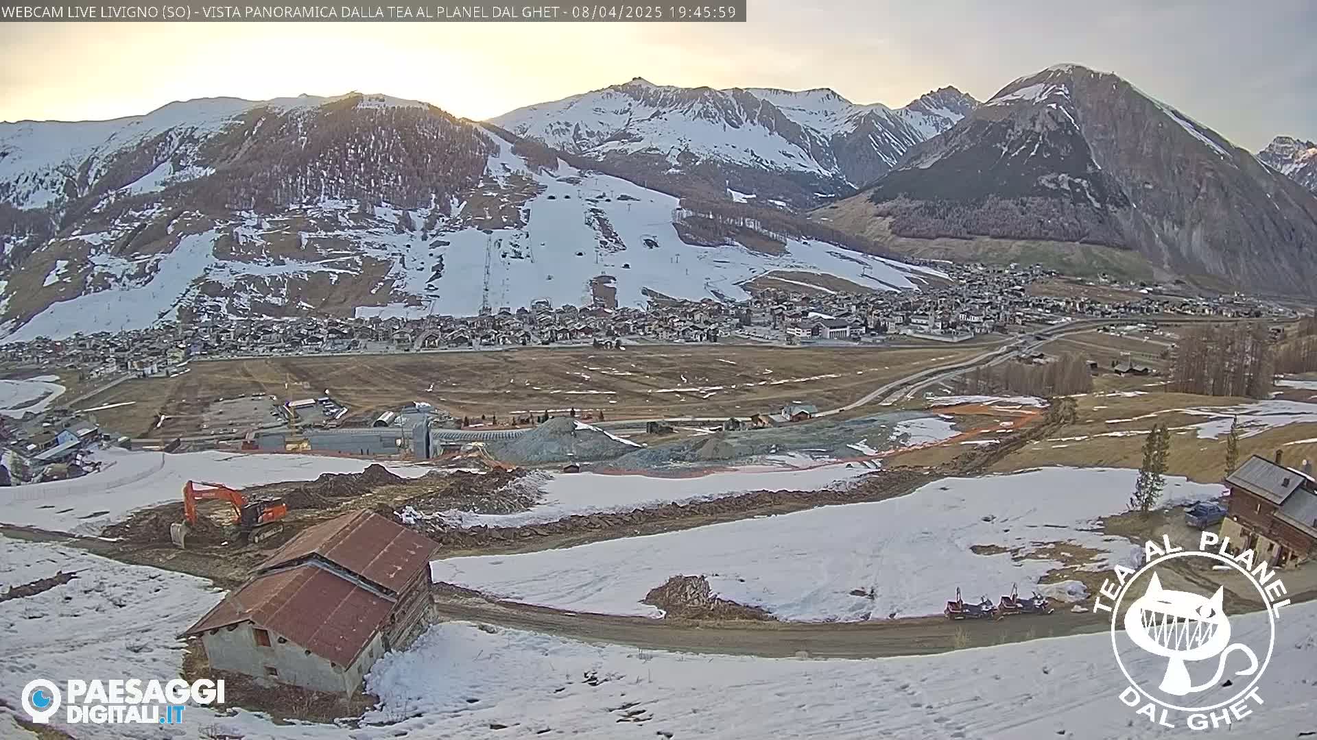 A partially snow-covered mountain village is seen from a hillside, with construction or demolition work taking place in the foreground under a partly cloudy sky.