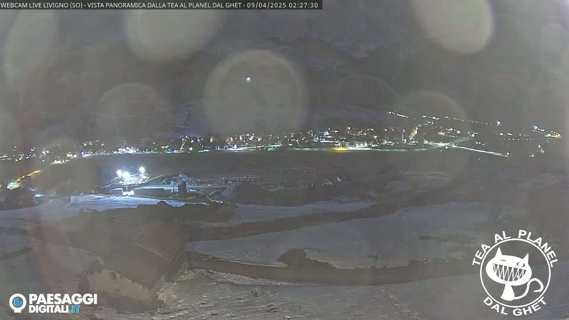 A nighttime, snowy landscape shows a town illuminated in the distance under a hazy, dark sky.