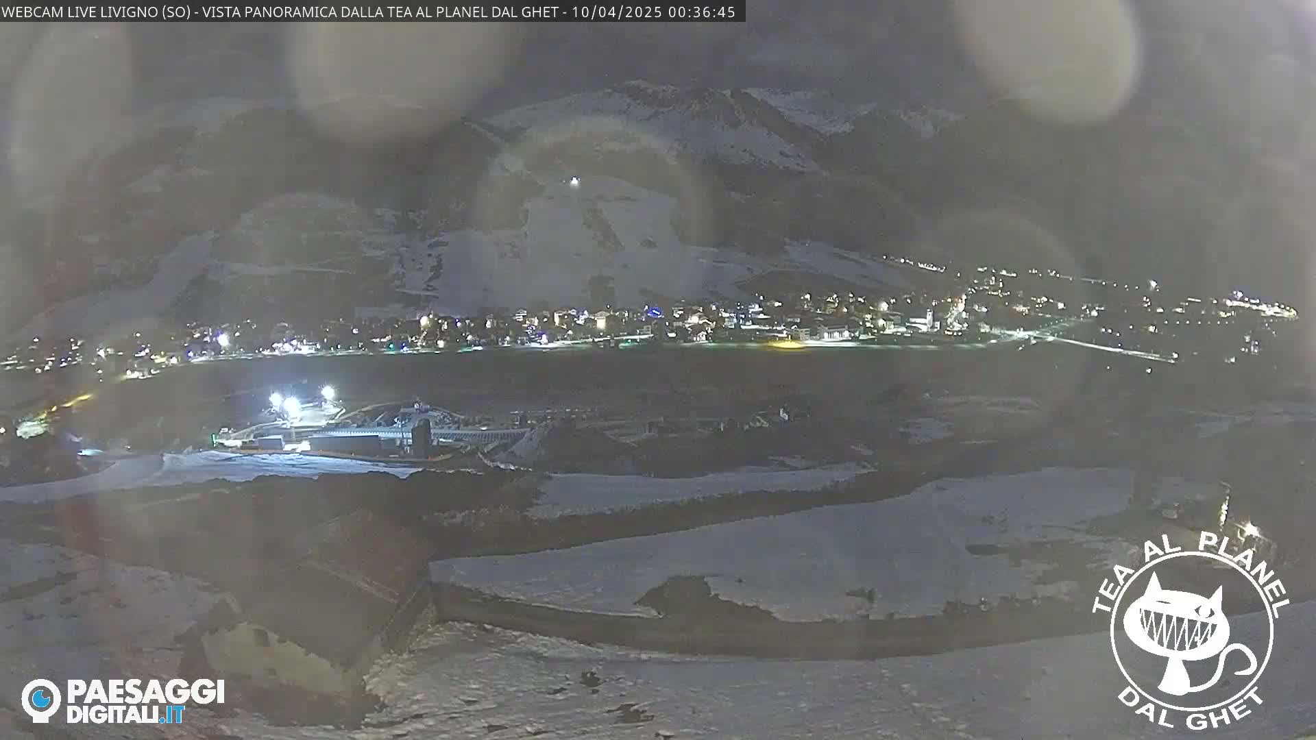 A nighttime, snowy, panoramic view of a town nestled in a valley, illuminated by lights, with snow-covered slopes in the foreground and background.