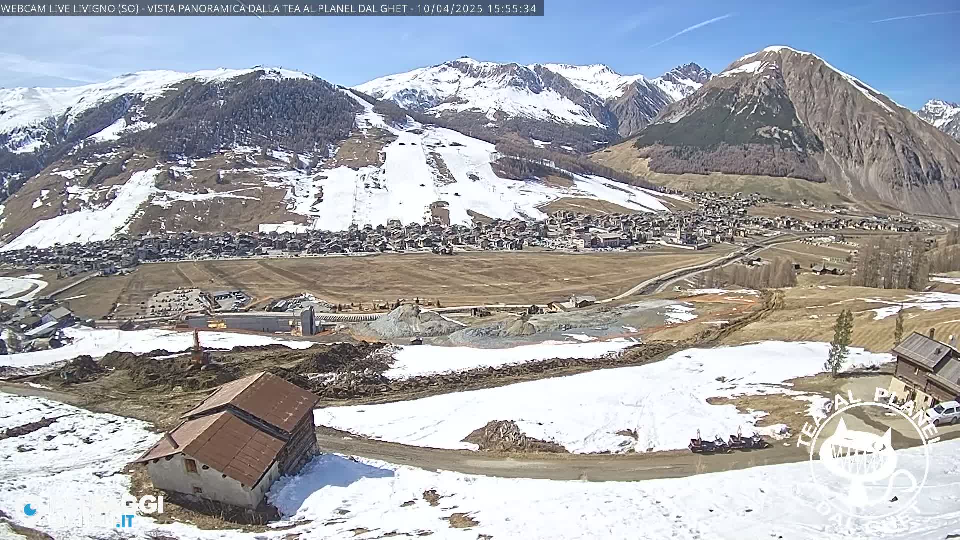 A partly snowy mountain village is visible under a sunny sky, with construction or land clearing visible in the foreground.