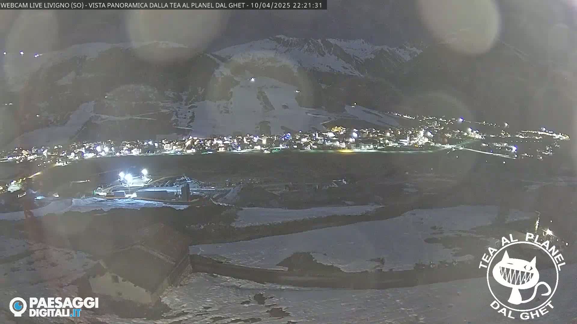 A nighttime, panoramic view of a snow-covered town nestled in a valley beneath snow-capped mountains.