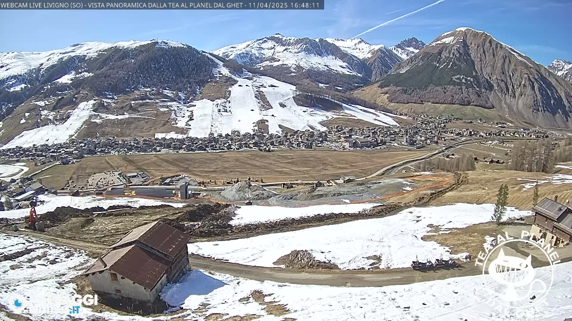 A partly snowy mountain landscape shows a village nestled in a valley below snow-capped mountains under a clear blue sky.