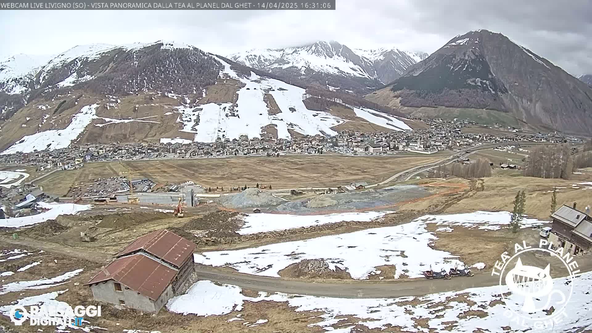 A partially snow-covered mountain town sits in a valley beneath snow-dusted mountains under a cloudy sky.