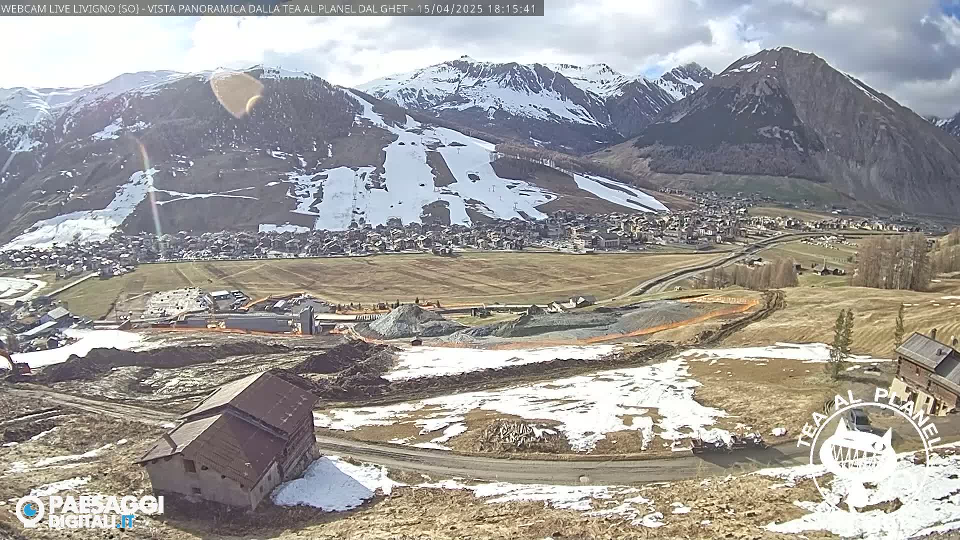 A partially snow-covered mountain valley with a town in the distance, under a mostly sunny sky, shows a construction site in the foreground with a tilted building.