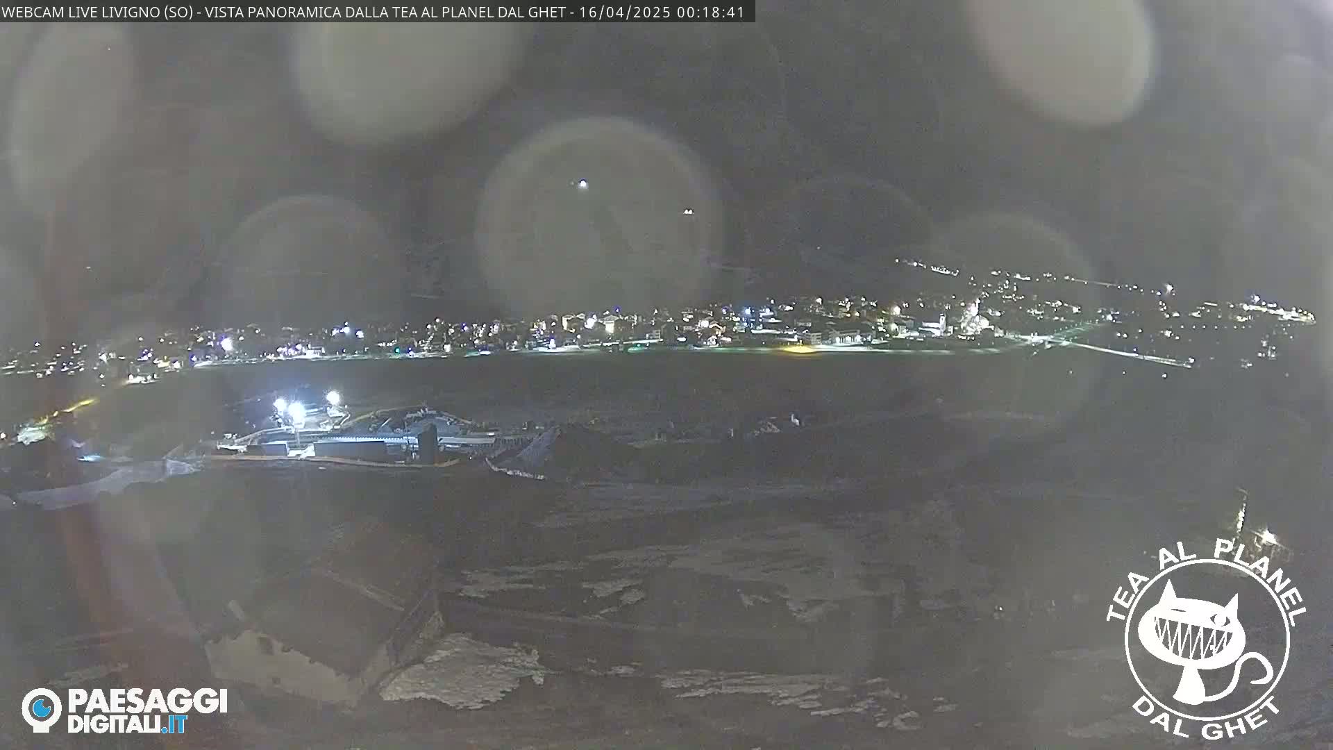 A nighttime panoramic view of a town illuminated by lights, with some snow visible in the foreground under a dark, clear sky.