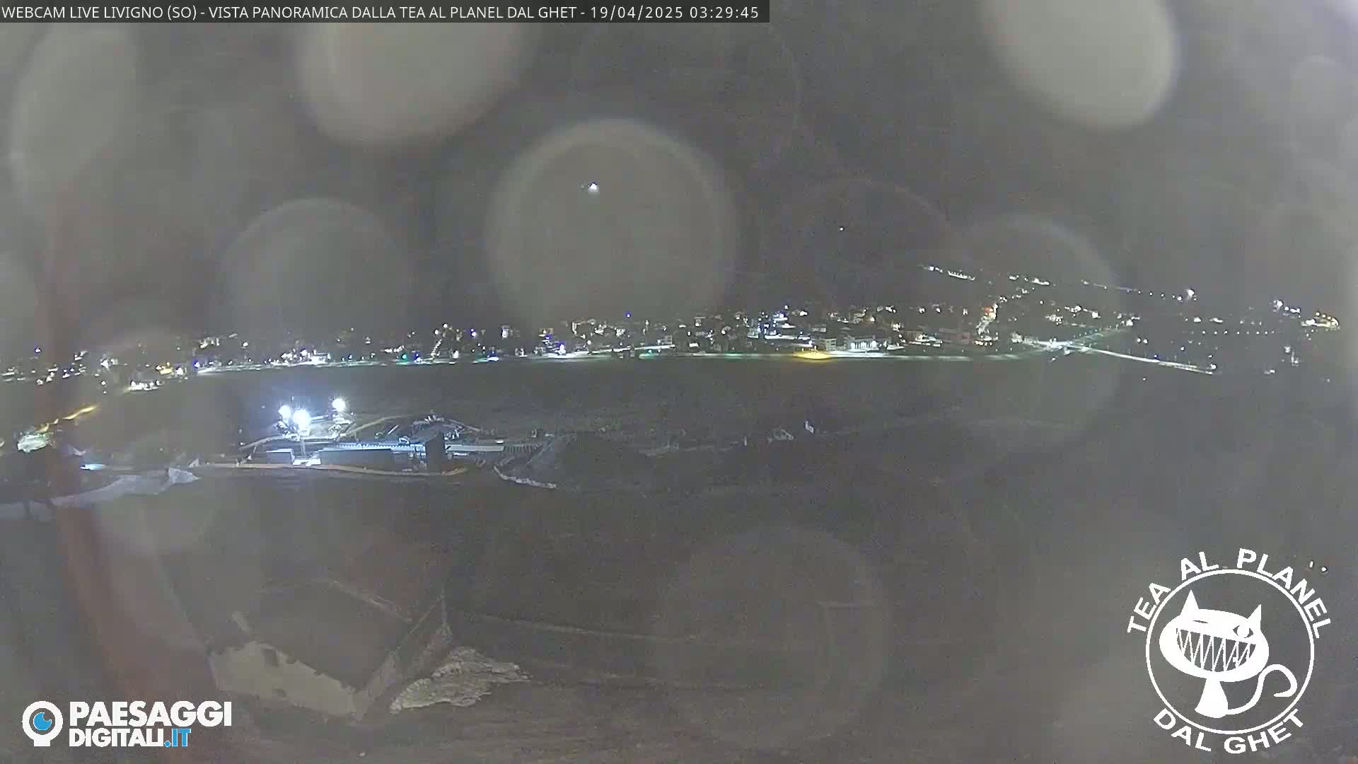 A nighttime, blurry, panoramic view of a snow-covered landscape showing a distant town with lights, under a dark, possibly cloudy sky.