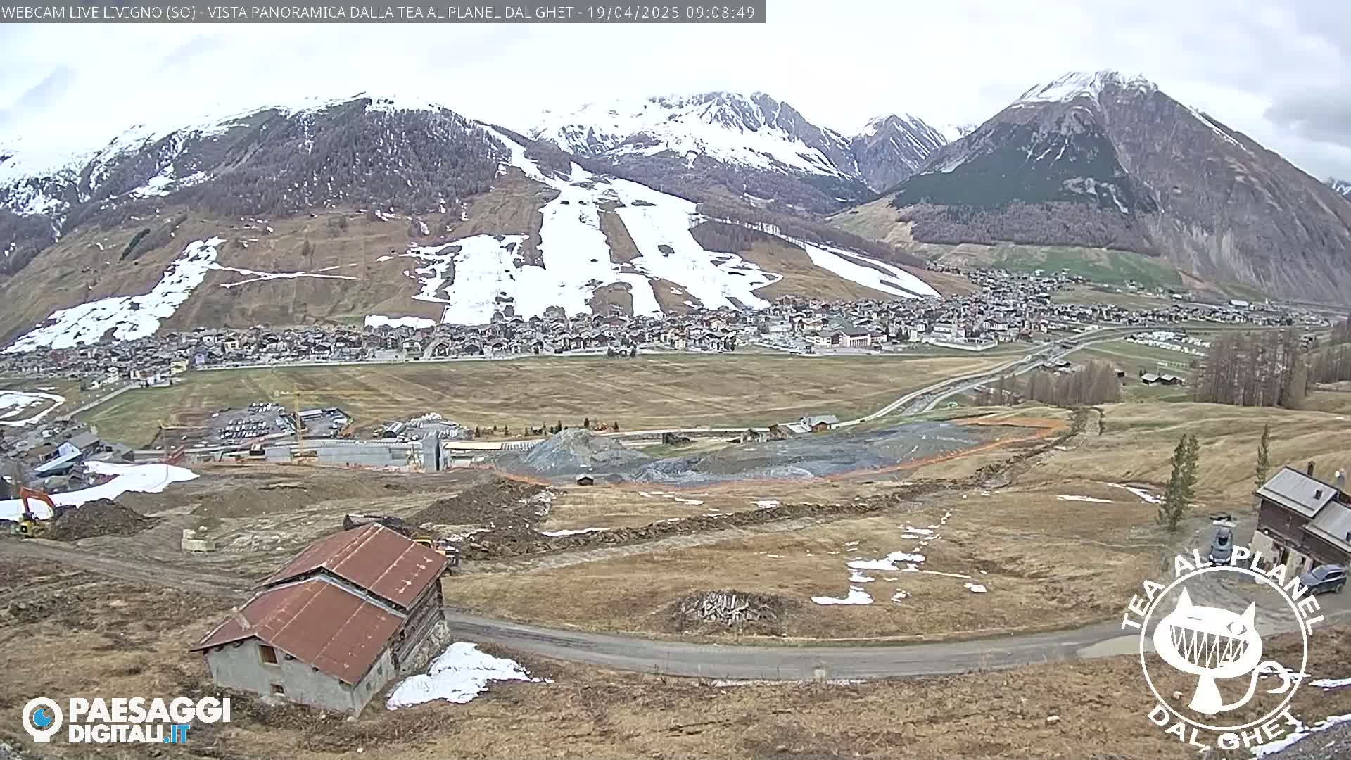 A long shot of a mountain town nestled in a valley below snow-capped mountains under an overcast sky, with a construction site in the foreground.