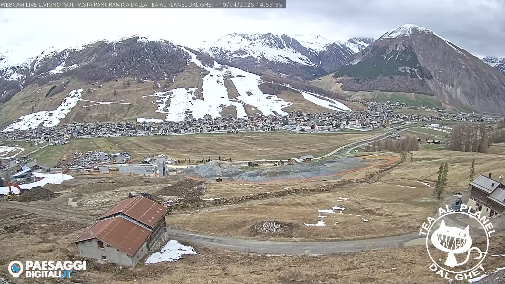 A long shot of a mountain town nestled in a valley, partially snow-covered mountains in the background, under an overcast sky, with construction visible in the foreground.
