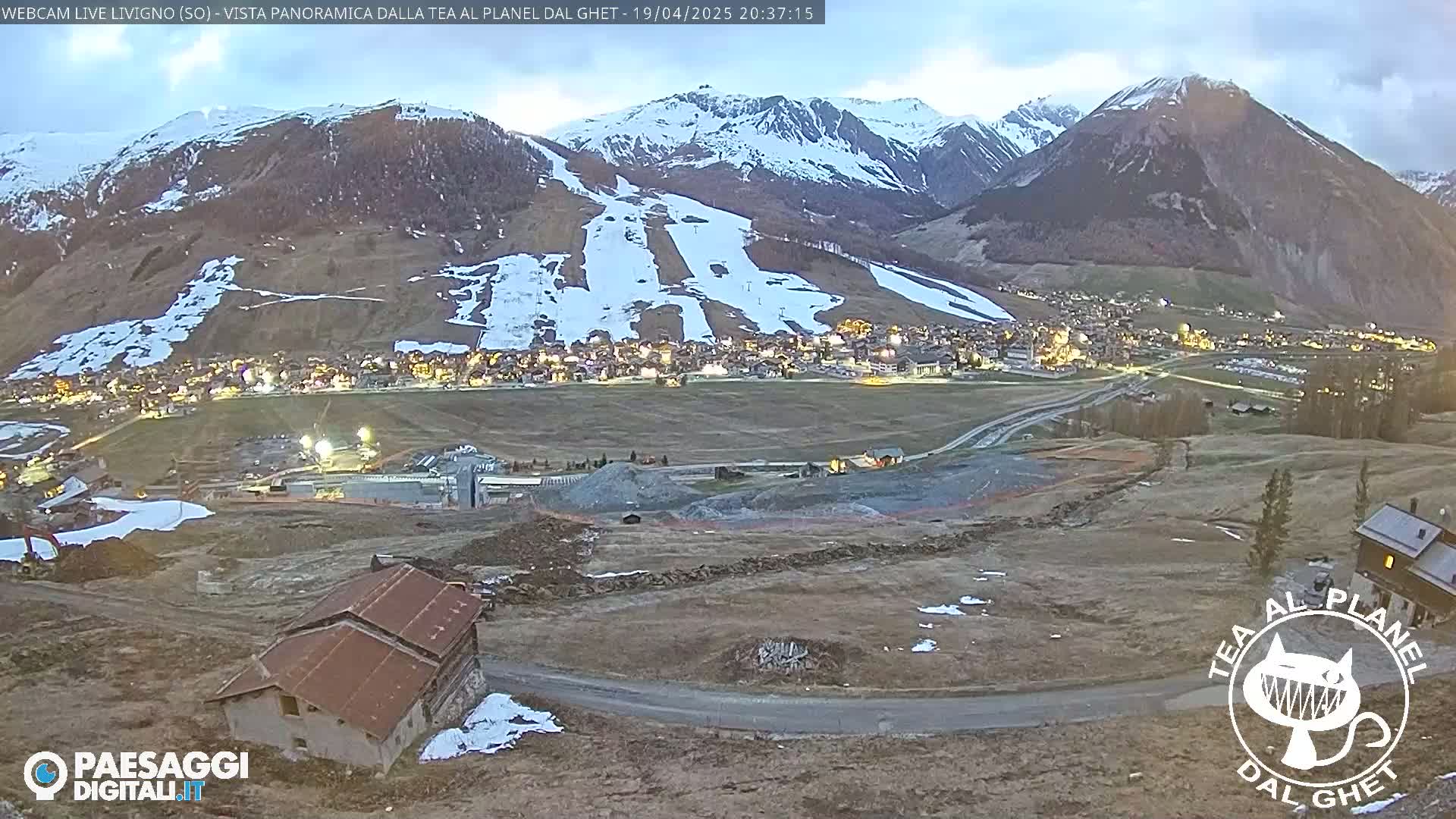 A mountain village at dusk is nestled below snow-dusted mountains under a twilight sky.