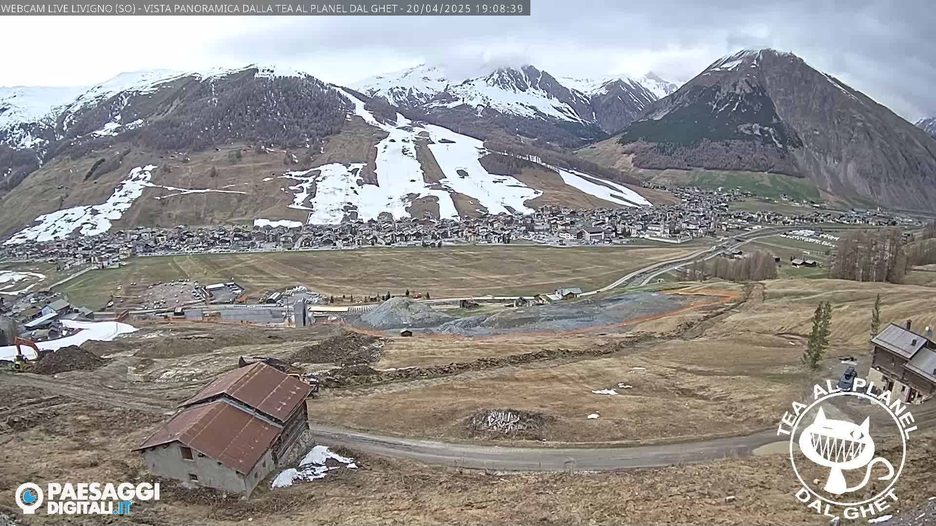 A partly cloudy view of a mountain town, partially snow-covered ski slopes, and a construction site, with a small building in the foreground.