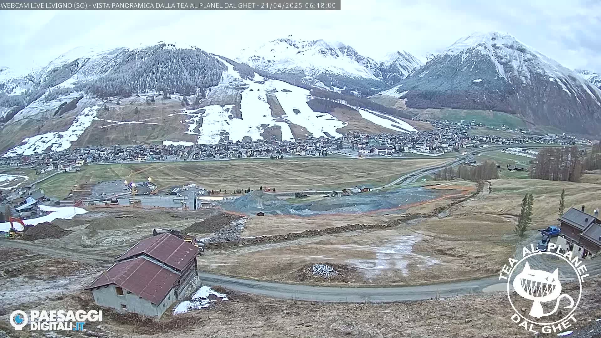 A snow-dusted mountain range overlooks a town and a construction site under an overcast sky.