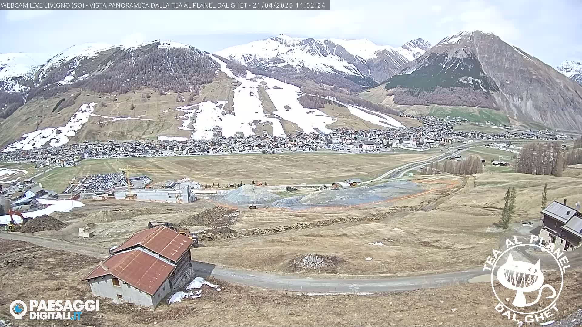 A partly sunny mountain town, with snow still on the slopes of the surrounding mountains, is seen from a high vantage point overlooking an area of construction.