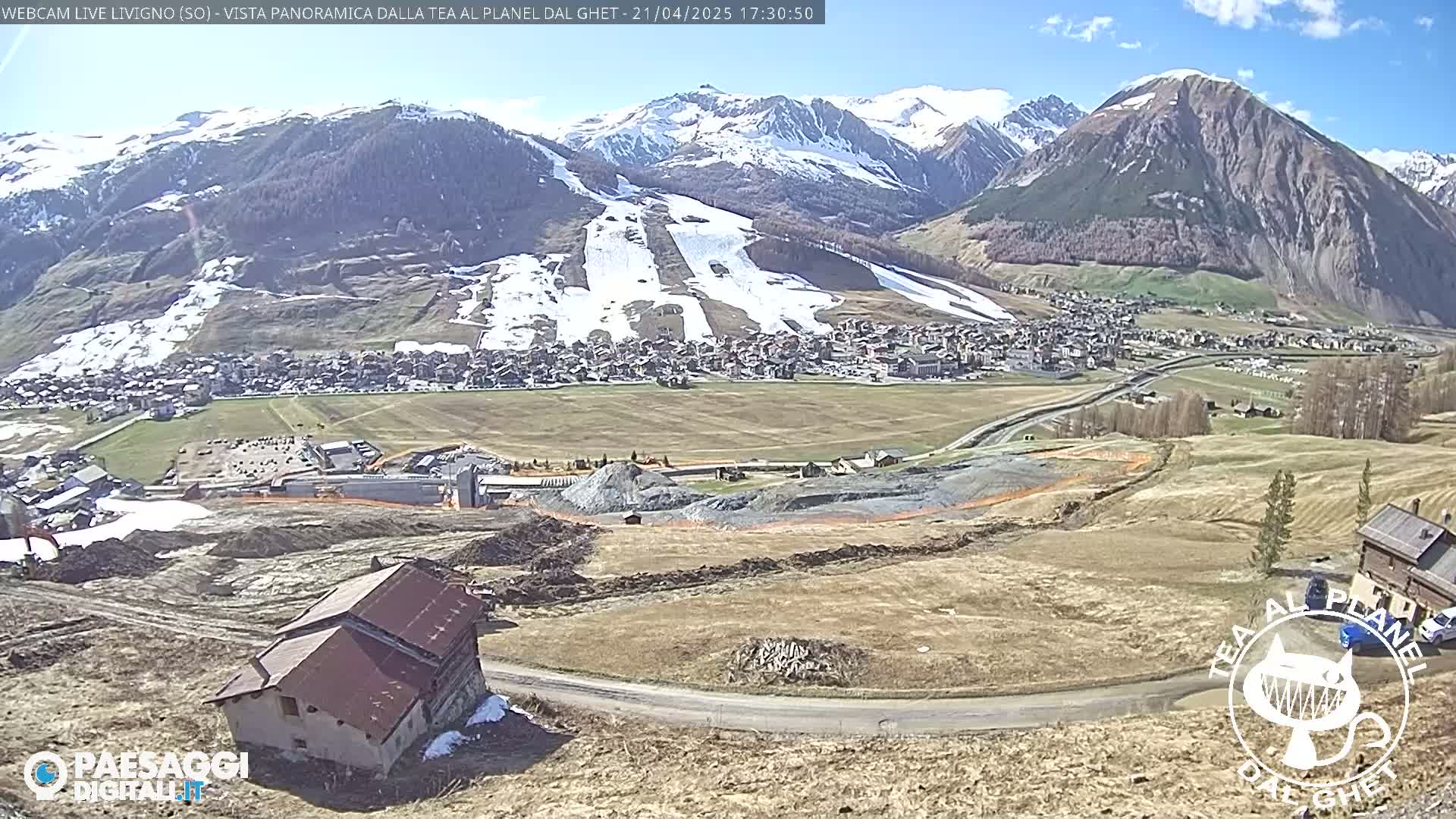 A mostly sunny, high-altitude view overlooking a village nestled in a valley, with snow-capped mountains in the background and a partially cleared construction site in the foreground.