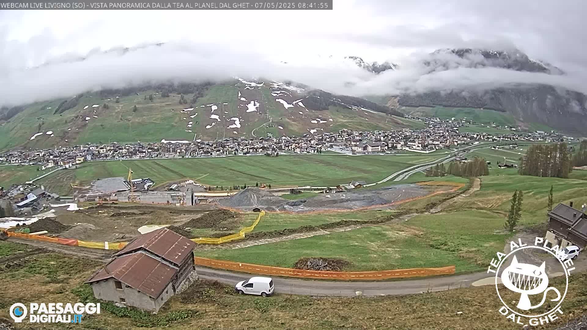 A cloudy day reveals a valley town nestled amongst green hills and mountains, partially obscured by fog, with construction visible in the foreground.
