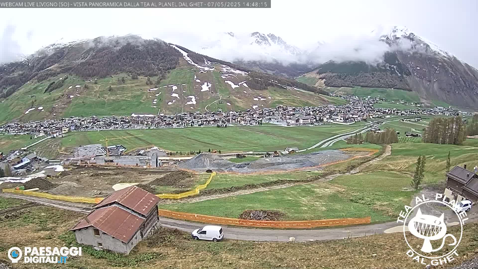 A partially cloudy day reveals a mountain village nestled in a valley, with a construction site and a white van visible in the foreground.